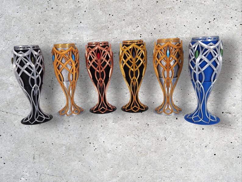 Six 3D-printed goblets in various colors, including silver, gold, copper, and blue, with intricate elven designs, are arranged in a row on a gray textured surface.