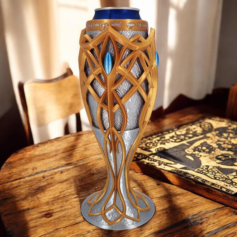 A gold and silver 3D-printed goblet, holding a blue soda can, sits on a wooden table next to an open book with intricate patterns.