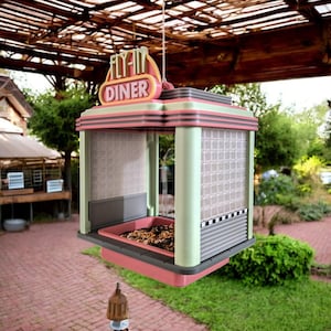 A pink, mint green, and gray retro diner-style bird feeder with "FLY-IN DINER" on its sign hangs from a string, filled with birdseed, with a brick patio in the background.