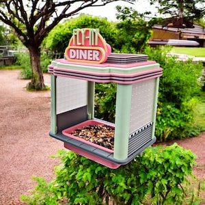 A pink, mint green, and gray retro diner-style bird feeder with "FLY-IN DINER" on its sign is suspended above a green bush, filled with birdseed.