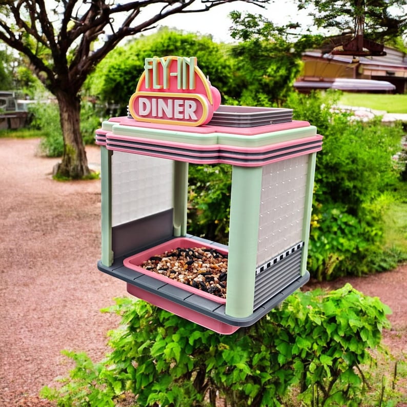 A pink, mint green, and gray retro diner-style bird feeder with "FLY-IN DINER" on its sign is suspended above a green bush, filled with birdseed.