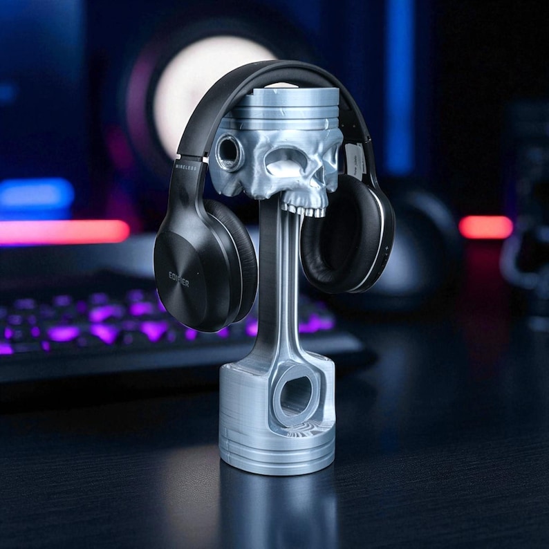 A silver-colored piston-shaped headphone stand with a skull head holds black wireless headphones on a dark desk with a glowing keyboard and mouse.