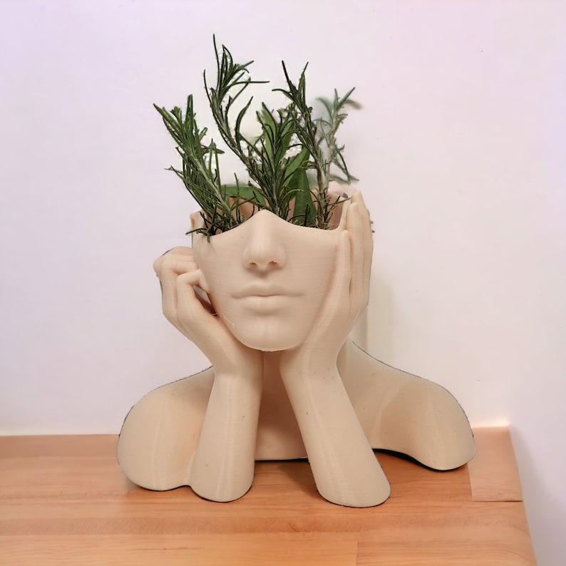 Op de afbeelding: Een witte keramische plantenpot in de vorm van een vrouwenhoofd met haar handen op haar kin. De pot heeft een gat in de bovenkant van het hoofd waar planten in kunnen worden geplaatst. De pot is gevuld met groene planten.