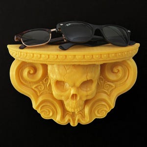 May include: A yellow wall-mounted shelf featuring a central skull carving surrounded by ornate scrollwork and a flat top surface. A pair of black-framed sunglasses with dark lenses rests on the shelf, positioned horizontally across the top.