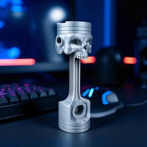 A silver-colored piston-shaped headphone stand with a skull head is angled on a dark desk, with a glowing keyboard and mouse in the background.