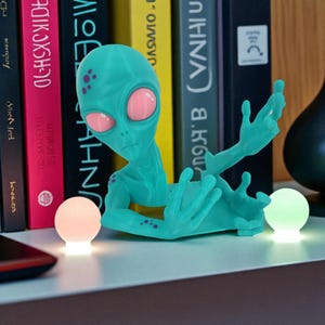 May include: A teal alien figurine with large pink eyes and purple spots on its head and arms. The alien is in a reclining pose with one arm raised. Two small, glowing spherical lights, one pink and one green, are positioned on either side.
