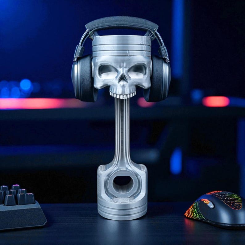 A silver-colored piston-shaped headphone stand with a skull head holds black wireless headphones, centered on a dark desk with a glowing keyboard and mouse.