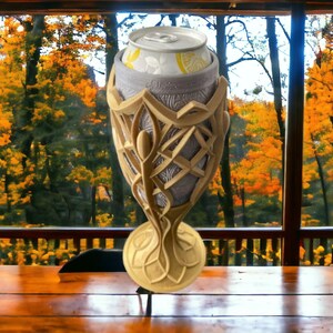 A gold and silver 3D-printed goblet, holding a white soda can with a lemon graphic, sits on a wooden table in front of a window with autumn trees.