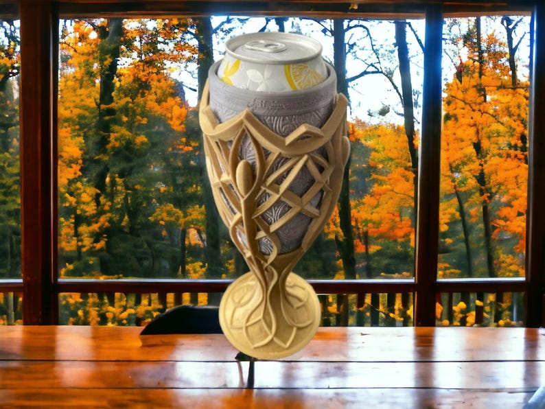 A gold and silver 3D-printed goblet, holding a white soda can with a lemon graphic, sits on a wooden table in front of a window with autumn trees.