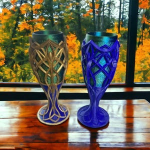 Two 3D-printed goblets, one gold and silver, the other blue, sit on a wooden table in front of a window with autumn trees.