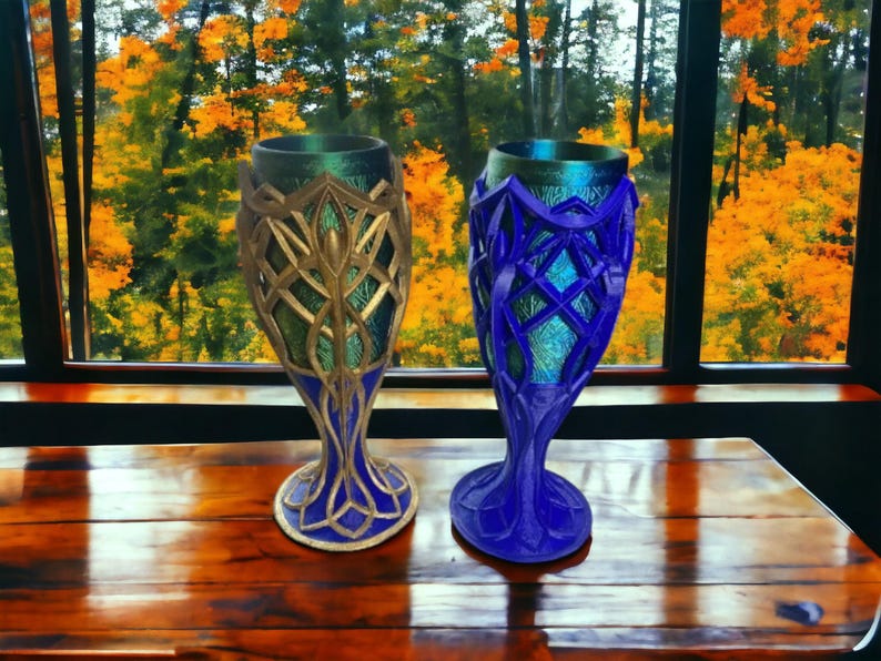 Two 3D-printed goblets, one gold and silver, the other blue, sit on a wooden table in front of a window with autumn trees.