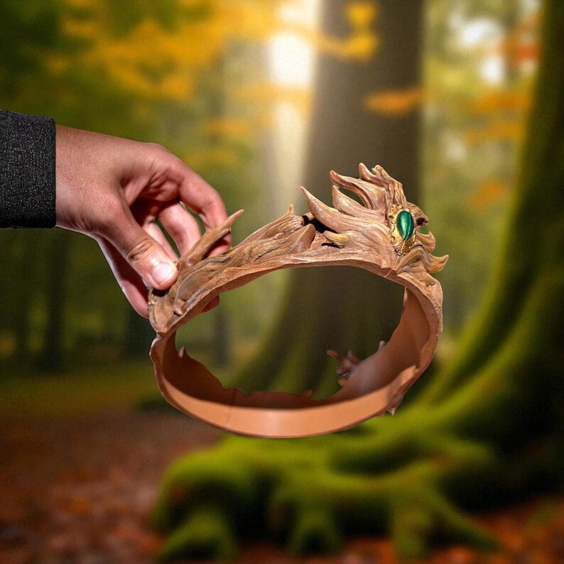 A hand holds a brown, textured crown with a green gem against a blurred forest background.