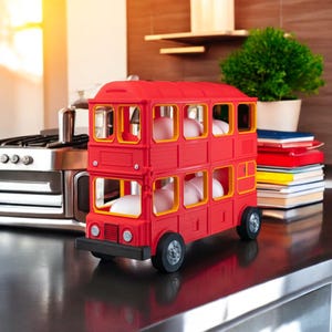 Peut inclure: Un support à œufs en forme de bus à impériale rouge avec des encadrements de fenêtres jaunes. Le bus jouet a des roues noires et est posé sur une surface sombre. Un empilement de livres et une petite plante en pot sont à côté du bus.