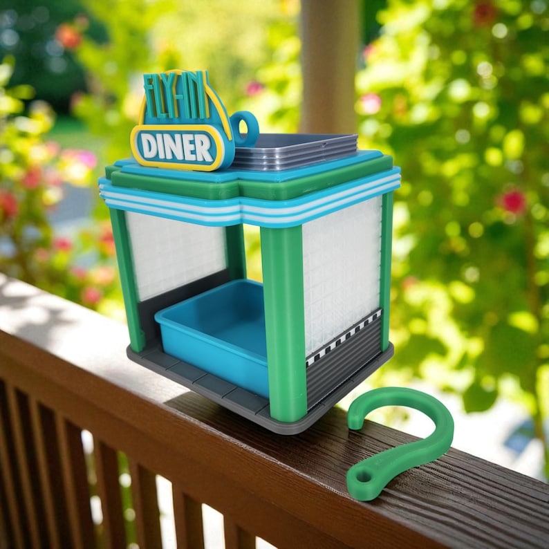 A blue, green, and gray retro diner-style bird feeder with "FLY-IN DINER" on its sign sits on a wooden railing next to a green plastic hook.