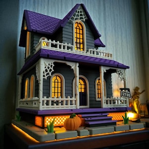 Puede incluir: Una casa en miniatura con temática de Halloween con exterior gris, techo morado y detalles blancos. La casa presenta ventanas iluminadas, un porche y telarañas decorativas. Dos calabazas naranjas están en los escalones. La casa tiene el número 2195 en una señal.