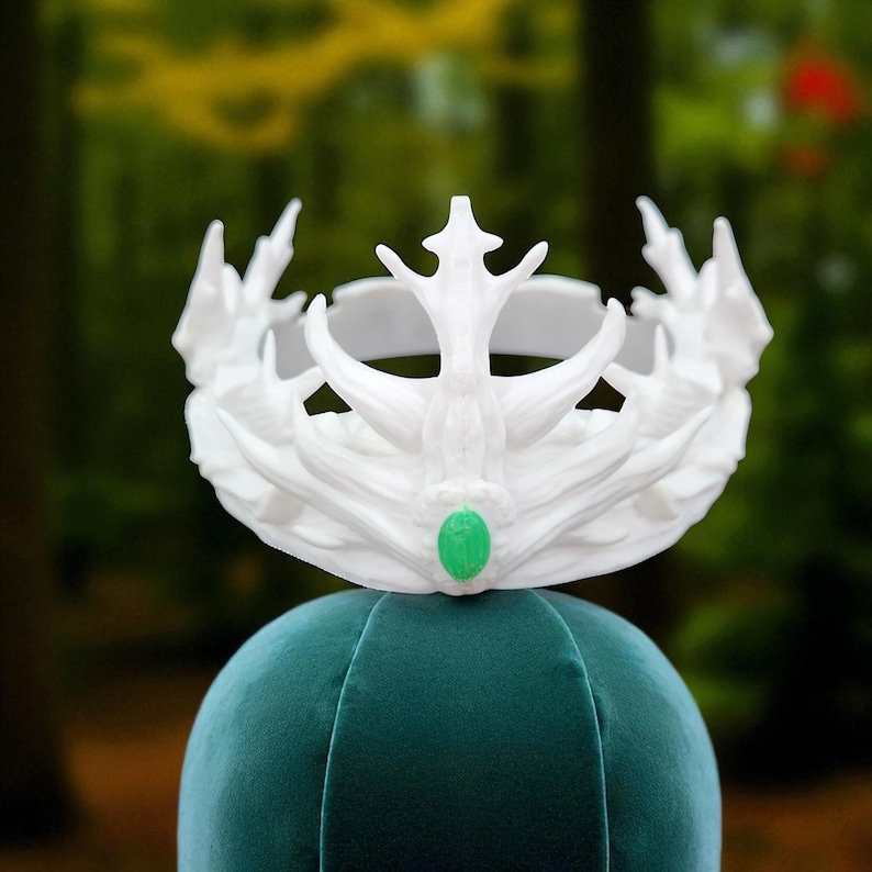 A white, textured crown with a green gem is displayed on a teal velvet head form against a blurred green background.