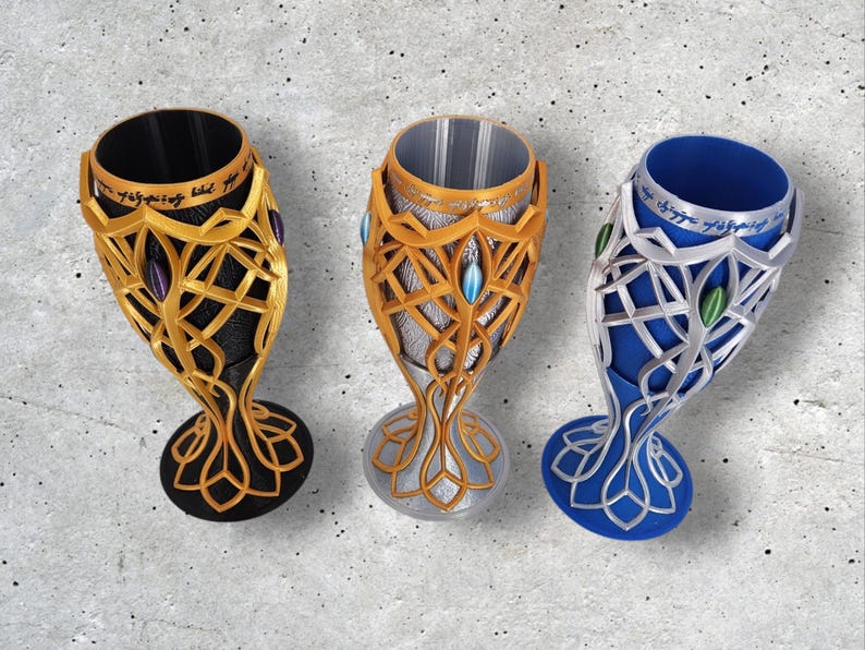 Three 3D-printed goblets in gold, silver, and blue, each with a black or silver interior and elven script, are displayed on a gray textured surface.