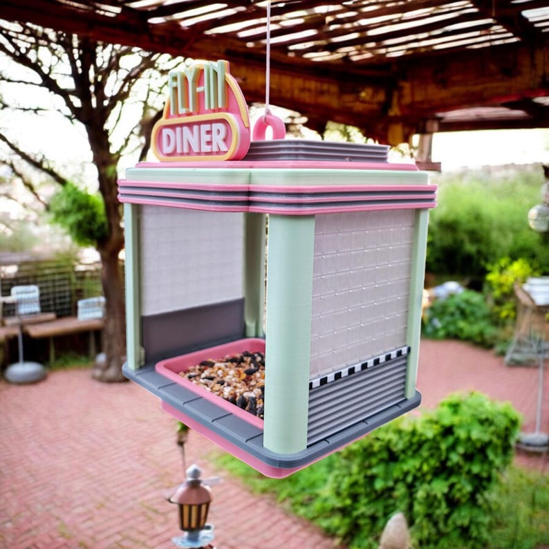 A pink, mint green, and gray bird feeder shaped like a retro diner, with "FLY-IN DINER" on top, hangs from a wooden pergola with birdseed in its tray.