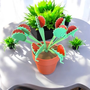 May include: A green plastic toy plant with red mouths that snap shut. The plant is in a terracotta pot and is surrounded by small green plants.