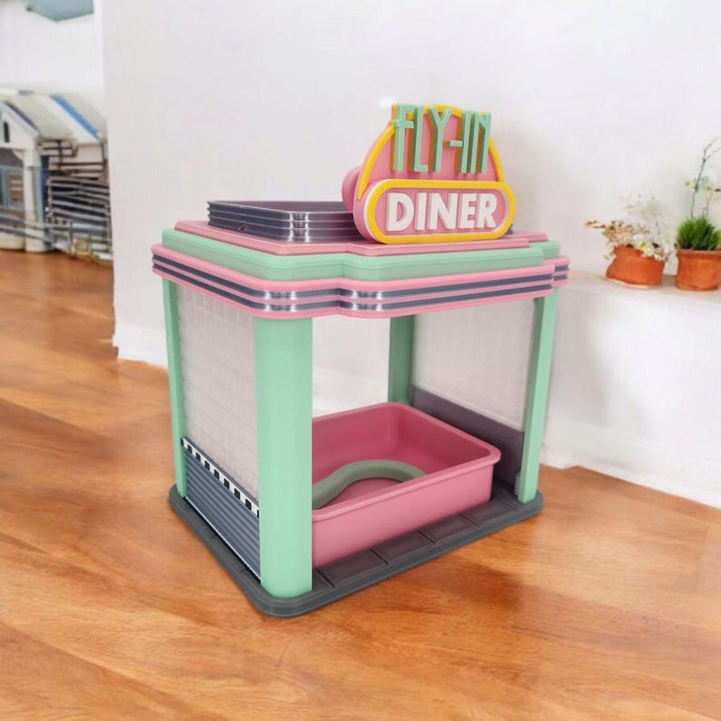 A pink, mint green, and gray retro diner-style bird feeder with "FLY-IN DINER" on its sign sits on a wooden floor against a white wall.