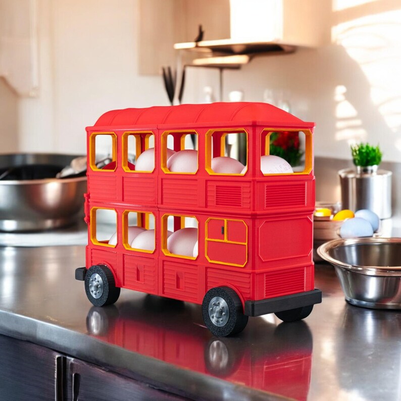 以下が含まれることがあります： 黄色い窓枠が付いた赤い二階建てバス型の卵ホルダー。 卵ホルダーには黒い車輪があり、白い卵で満たされています。 バスは、キッチンのステンレス鋼のカウンタートップに置かれています。