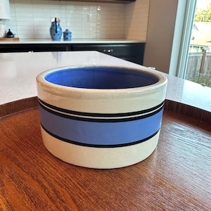 May include: A cylindrical ceramic bowl with a cream-colored exterior featuring a wide horizontal light blue band bordered by two thin black stripes. The interior of the bowl is a solid, deep blue color, and the rim is a smooth, rounded edge.