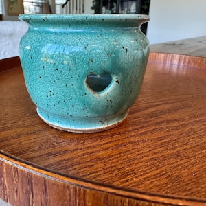 May include: A teal ceramic pot with a speckled finish and a small heart-shaped cutout on the front. The pot has a rounded body, a flared rim, and a visible unglazed base. The surface texture appears smooth with dark brown speckles throughout.