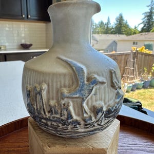May include: A beige ceramic pitcher featuring a relief design of flying birds and waves in blue and dark gray tones. The vessel has a rounded body, a narrow neck, and a flared rim, with a textured surface that highlights the embossed nautical imagery.