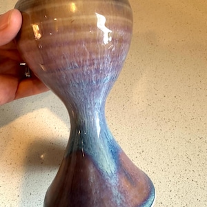 May include: A ceramic goblet featuring a flared rim and a narrow stem that widens into a circular base. The glaze displays a gradient of colors, transitioning from a warm tan at the top to a deep blue and purple hue toward the bottom of the vessel.