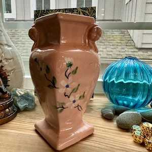 May include: A pale pink ceramic vase with a square base and rim, featuring two small sculpted cherub-like figures on the upper sides. The front of the vase is decorated with a hand-painted floral motif consisting of white blossoms with blue centers.