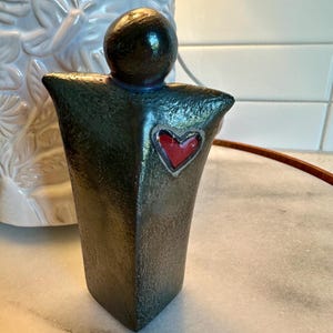 May include: A dark green, textured ceramic figurine with a smooth, rounded head and a triangular body. The figure features a small, glossy red heart shape embedded in the center of the chest area, creating a distinct contrast against the dark finish.