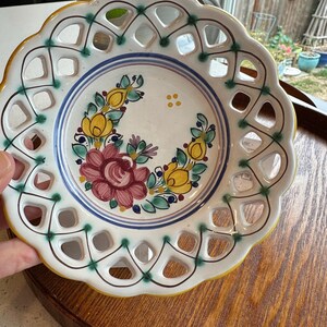 May include: A round ceramic bowl with a scalloped edge and an intricate lattice-cut border. The center features a hand-painted floral motif with a large red rose, yellow buds, and green leaves, accented by blue circular lines and green dots.