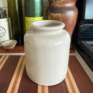 May include: A cylindrical ceramic jar with a smooth, off-white glossy glaze. The vessel features a wide, rounded rim and a straight-sided body that tapers slightly toward the base. The surface is uniform in color with a subtle, reflective finish.