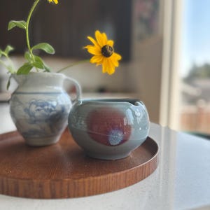 May include: A small, round, light blue ceramic bowl with a prominent reddish-brown circular glaze pattern on its side, positioned next to a white ceramic pitcher featuring a blue floral design and two yellow flowers with dark centers.