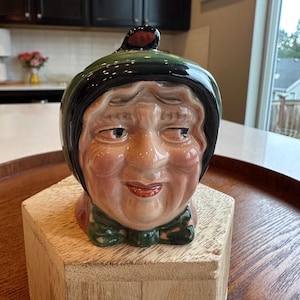 May include: A ceramic character jug shaped like an elderly person's head, wearing a dark green bonnet with a black trim and a matching green bow at the neck. The face features detailed facial expressions, pinkish skin tones, and painted eyes.