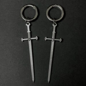 Pode incluir: Um par de brincos de espada prateados com um design de aro. As espadas têm uma ponta pontiaguda e uma guarda.