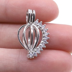 May include: Silver charm with a cage design and a half-circle of clear cubic zirconia stones. The charm is designed to hold a pearl or other small object.