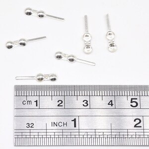 May include: Five silver-toned metal bead caps with a straight pin. The bead caps are approximately 1 inch long and have two round beads. The bead caps are shown on a ruler with inch and centimeter markings.