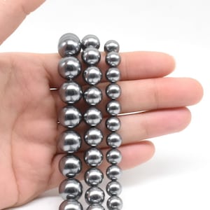 May include: Strands of gray faux pearls in varying sizes. The pearls are a uniform gray color and have a smooth, glossy finish. The pearls are held in a hand against a white background. The pearls are likely for jewelry making.