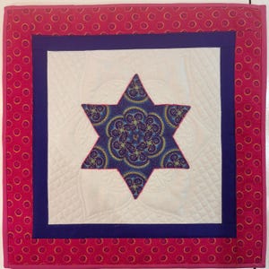 May include: A square quilted wall hanging with a central six-pointed star design. The star is a deep blue with a swirling pattern, set against a white background. The quilt has a dark blue border, and a pink border with yellow circles.