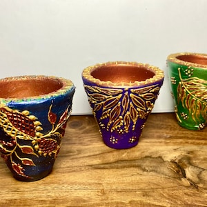 Könnte beinhalten: Drei dekorative, handbemalte Keramiktöpfe in Blau, Lila und Grün. Jeder Topf hat einen metallisch goldenen Rand und aufwendige goldene Blumendesigns. Die Töpfe werden auf einer Holzoberfläche ausgestellt.