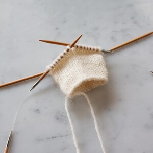 May include: A small piece of off-white knitted fabric held on circular knitting needles. The project features a stockinette stitch pattern with visible loops on the wooden needle tips and a flexible clear cord connecting the needle ends.