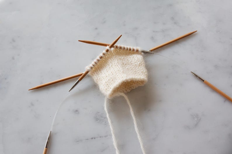 May include: A small piece of off-white knitted fabric held on circular knitting needles. The project features a stockinette stitch pattern with visible loops on the wooden needle tips and a flexible clear cord connecting the needle ends.