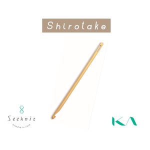 Könnte beinhalten: Ein hellfarbiger Häkelhaken aus Holz mit einem Haken an einem Ende und einem kleinen Loch am anderen. Das Wort "Shirotake" steht über dem Haken. Das Seeknit-Logo befindet sich in der unteren linken Ecke und das KA-Logo in der unteren rechten Ecke.