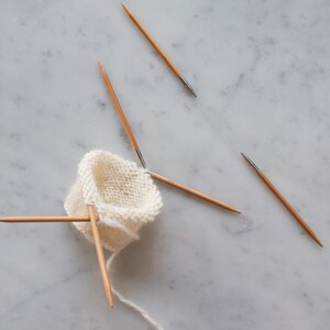 May include: A small, circular piece of off-white knitted fabric held by four thin, light brown wooden double-pointed knitting needles. The needles are arranged in a cross-like formation, with one needle extending downward with a trailing yarn tail.