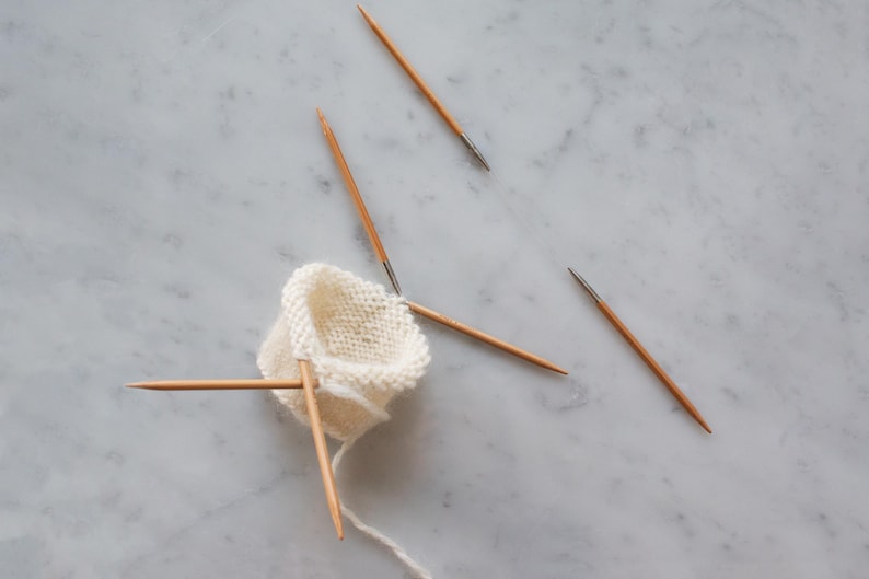 May include: A small, circular piece of off-white knitted fabric held by four thin, light brown wooden double-pointed knitting needles. The needles are arranged in a cross-like formation, with one needle extending downward with a trailing yarn tail.