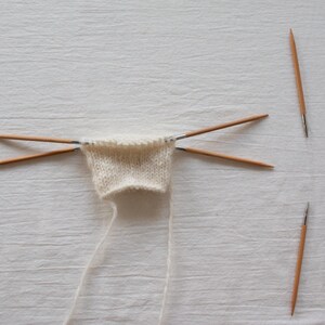 May include: A small piece of off-white knitted fabric held by four thin wooden double-pointed knitting needles arranged in a cross shape, with two additional matching wooden knitting needles placed separately to the right side of the fabric.