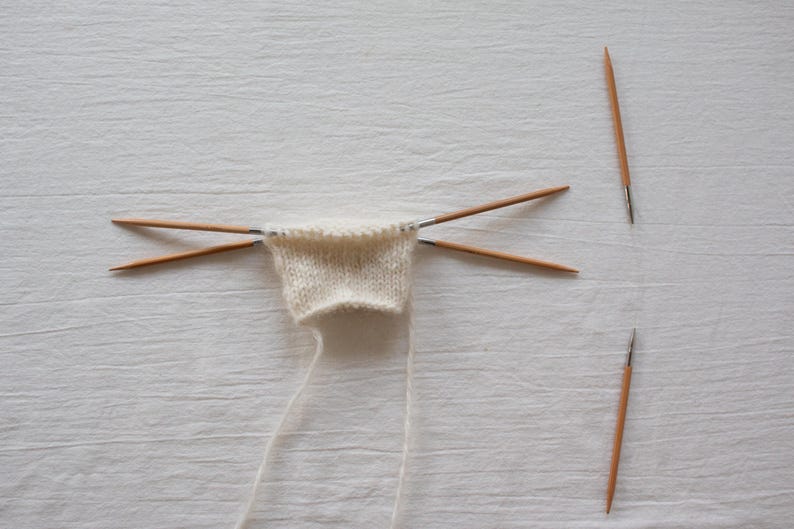 May include: A small piece of off-white knitted fabric held by four thin wooden double-pointed knitting needles arranged in a cross shape, with two additional matching wooden knitting needles placed separately to the right side of the fabric.