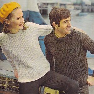 May include: A couple wearing matching cable knit sweaters. The woman is wearing a yellow beret and black pants. The man is wearing brown pants. They are standing in front of a wooden boat.