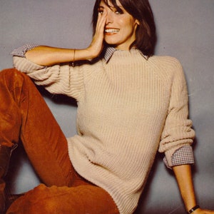 May include: A woman wearing a cream-colored knitted sweater with a brown and white plaid shirt collar peeking out. She is wearing brown corduroy pants and a gold bracelet on her wrist.